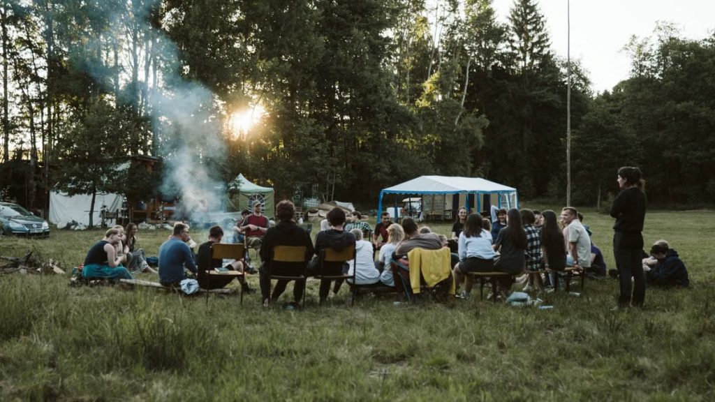A group of adults relaxing around a campfire in a scenic outdoor setting with tents.